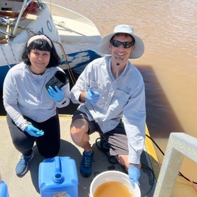 Two people on a dock collecting water samples with a boat and brown river behind them.